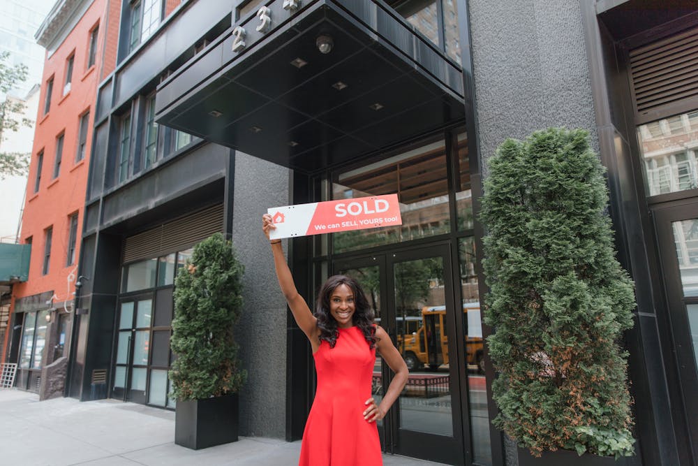 a woman wearing a red dress holding a sold sign in front of a building
